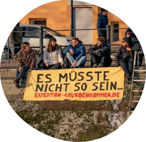 5 Mitglieder der Expedition Grundeinkommen sitzen zusammen und halten ein Transparent, auf dem "Es müsste nicht so sein.... Expedition-grundeinkommen.de" steht