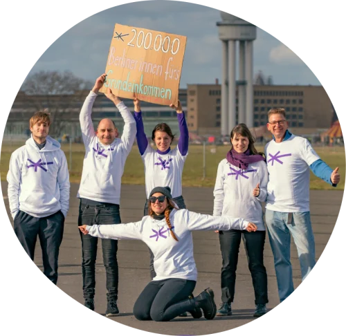 Die Mitglieder der Expedition Grundeinkommen stehen auf dem Tempelhofer Feld und halten ein Plakat mit der Aufschrift "200.000 Berliner innen fürs bedingungslose Grundeinkommen