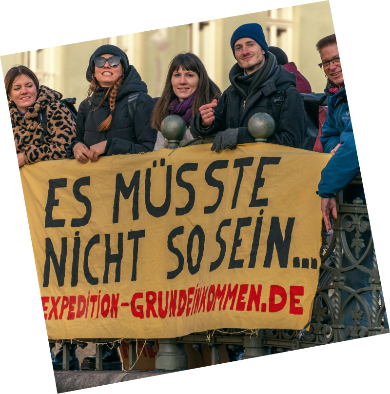Eine Gruppe von Menschen aus der Expedition mit einem Transparent mit der Aufschrift "Es müsste nicht so sein... www.expedition-grundeinkommen.de"